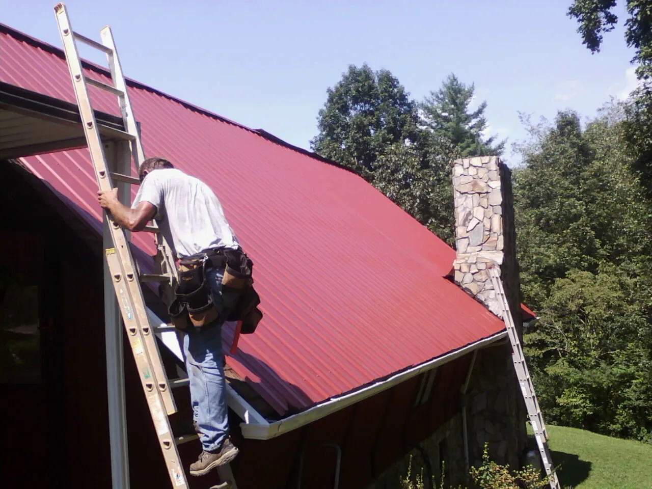 Roofer on a ladder beside a red metal roof and stone chimney — Soffit & Fascia Repair by Roofing Brothers in Dearborn Heights